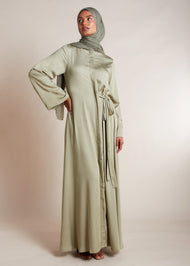 Discover the elegant and versatile Side Tie Abaya in an exquisite soft sage colour, crafted from a luxurious cotton satin blend that provides a subtle sheen. This abaya features a unique attached side belt that adds shape and dimension to the garment. 